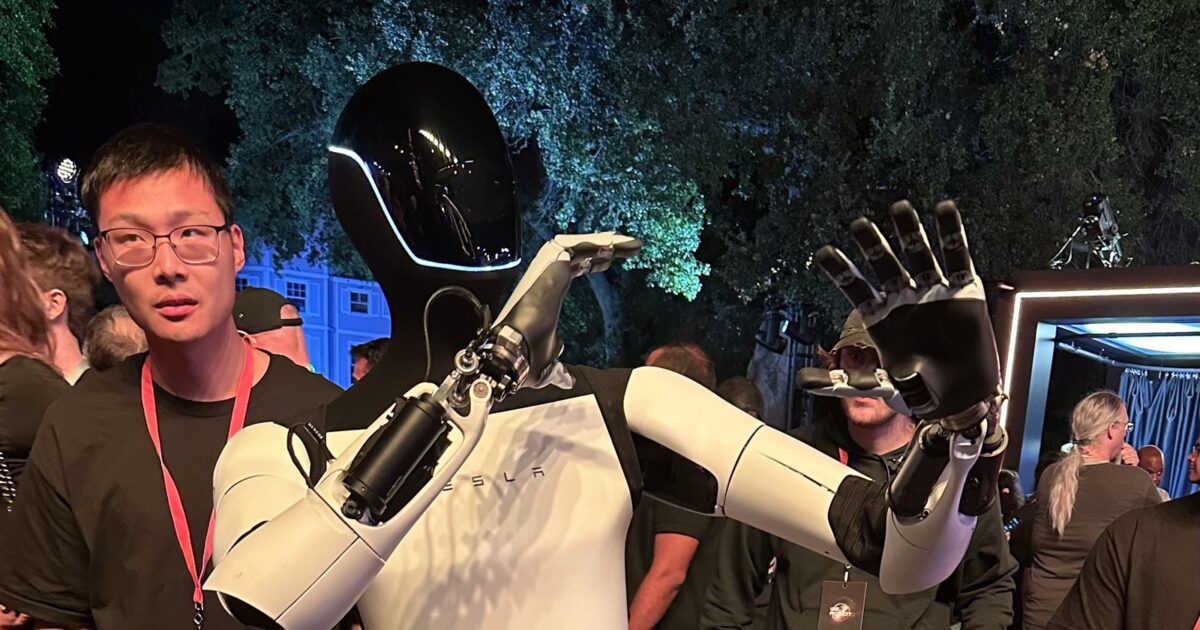 Tesla Optimus V3 humanoid robot waves to a crowd, showing off its dexterous hand and forearm design at a Tesla event.