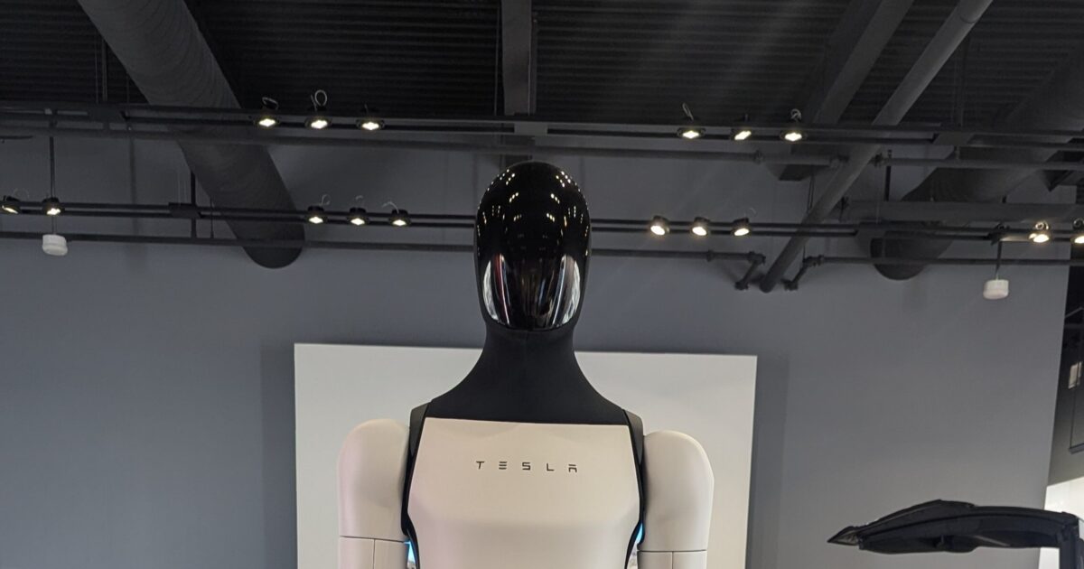 Tesla Optimus V3 humanoid robot on display in a Tesla showroom, showing the sleek white body panels and glossy black visor head with TESLA badge on the chest
