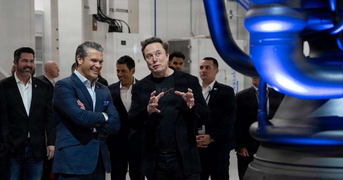 Elon Musk speaks beside visitors and aerospace hardware inside a SpaceX facility